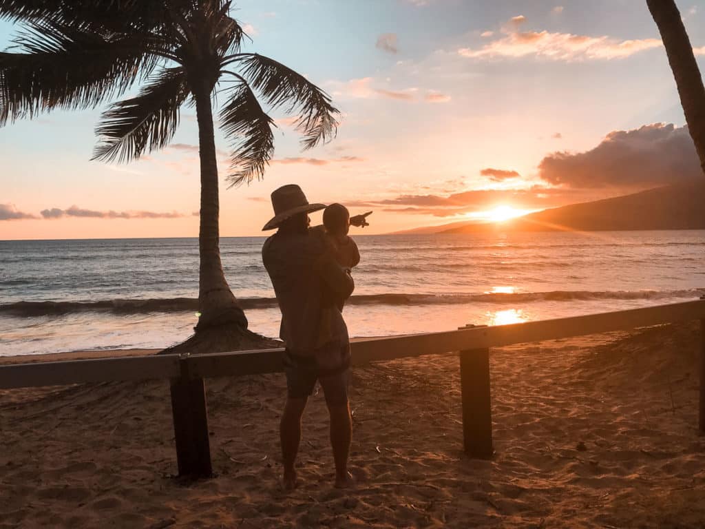 Dad pointing at sunset while looking out at the ocean and holding baby in Maui, Hawaii.