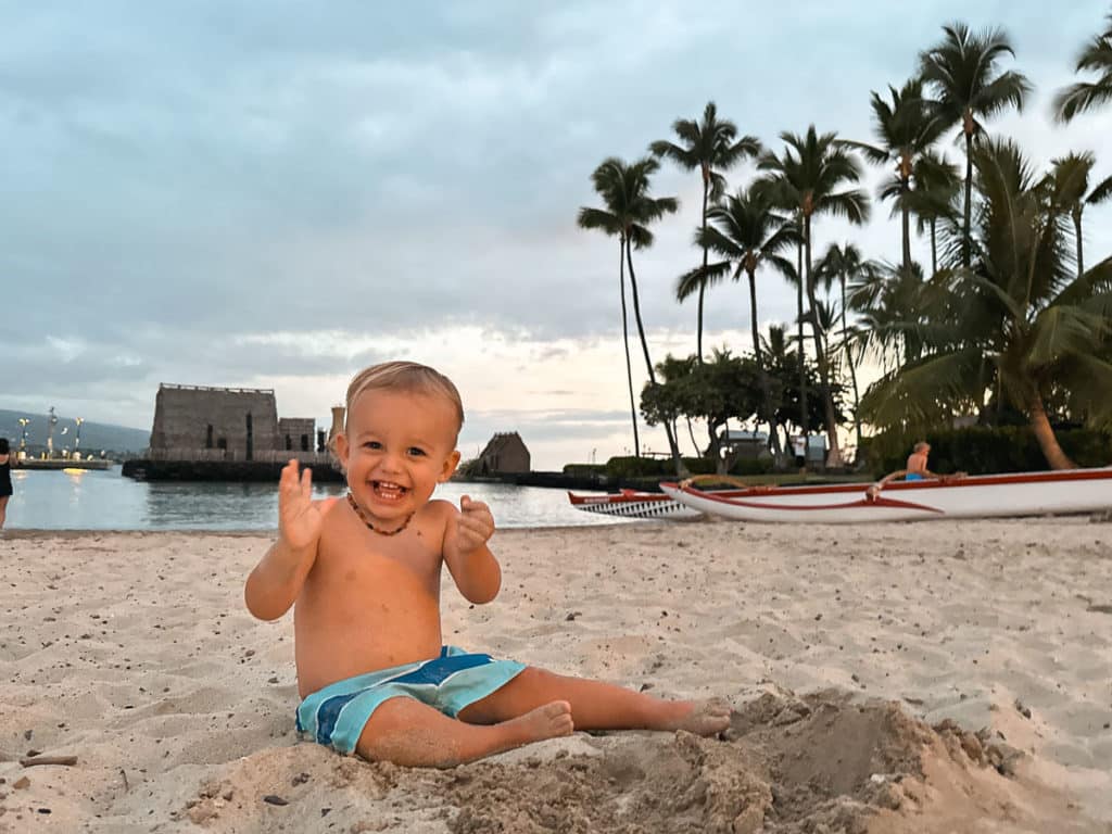 A toddler smiling and playing in the sand in front of the protected cove at King Kamehameha 's Kona Beach Hotel.