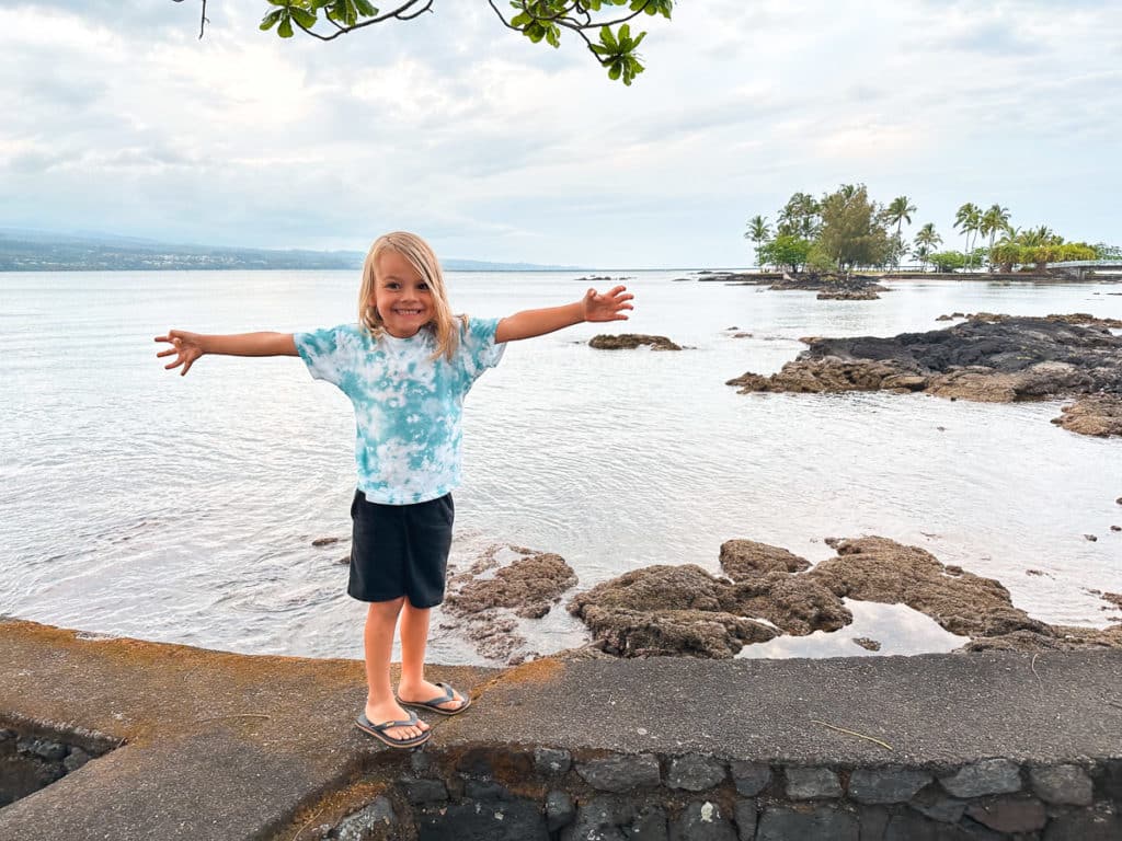 A boy with long hair smiling and standing with his arms outstretched at Coconut Island, Big Island, Hawaii. 