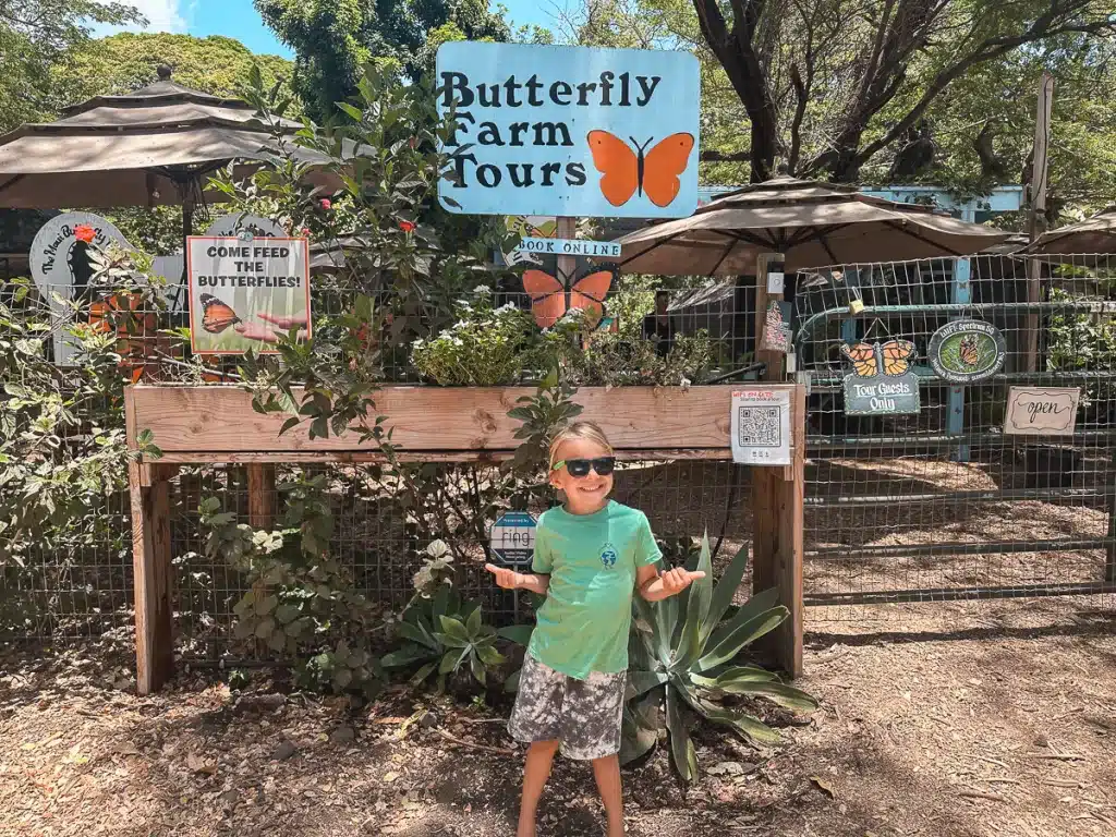 A young boy stands in front of the Maui Butterfly Farm sign in Maui, Hawaii. 