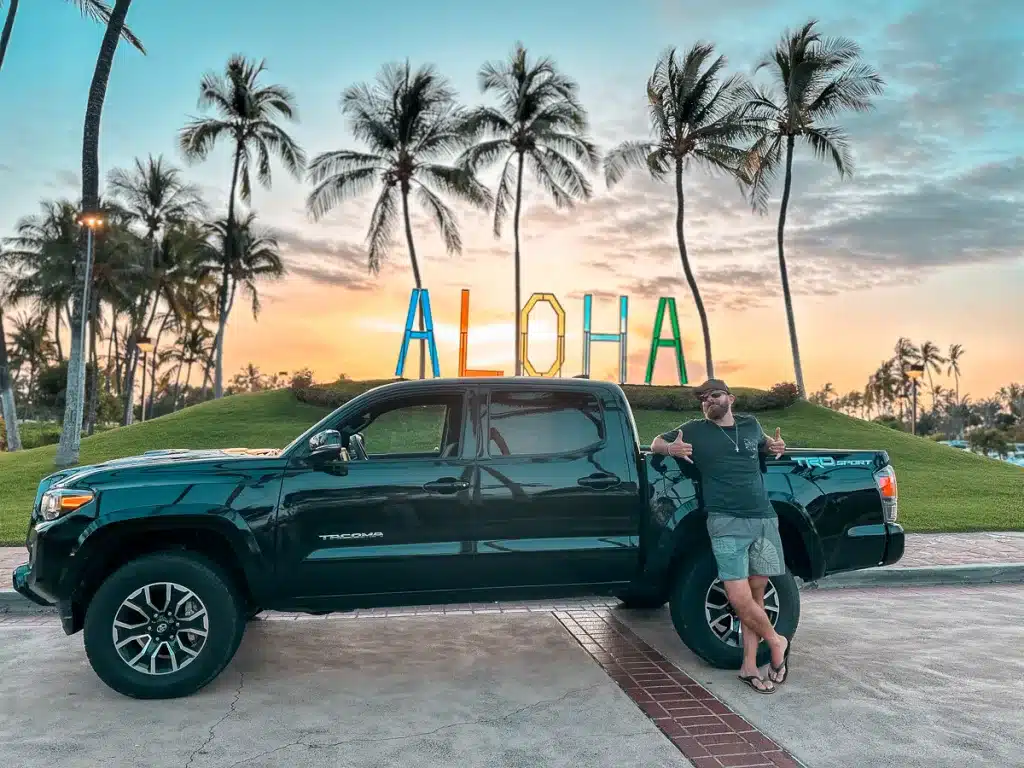 A man giving shakas and standing in front of a black truck and an aloha sign on the Big Island of Hawaii.