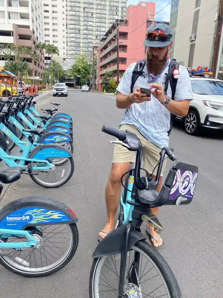 A man on a bicycle in Waikiki, looking at directions on his phone with buildings in the background. 