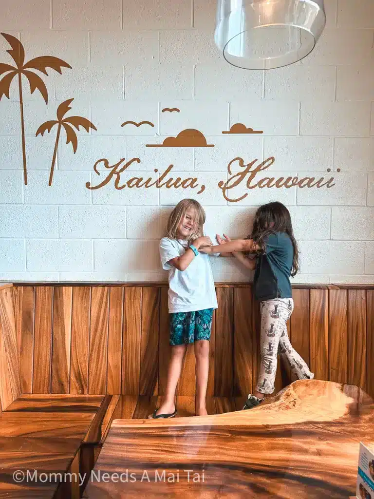 Two kids tickling each other and laughing under a painted wall that reads "Kailua, Hawaii."