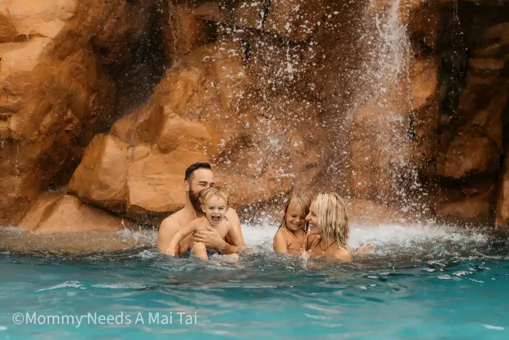 A family of four laughing and playing under a waterfall in Hawaii. 