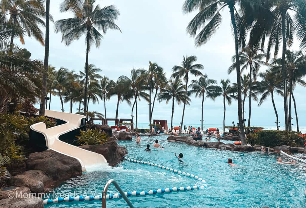 A view of the waterslide pool at Sheraton Waikiki, with kid's playing, palm trees, and the blue ocean in the background.