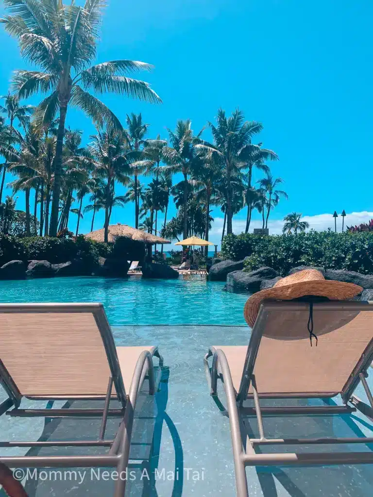 A sun hat resting on a chair in front of a resort pool with palm trees in the distance in Maui, Hawaii. 