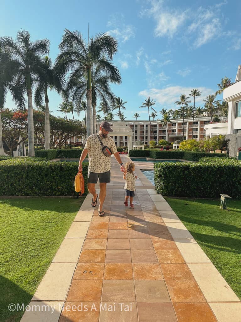 A dad holding hands with a toddler in a matching Aloha Shirt with the resort in the backgrond at the Grand Wailea Resort in Wailea, Maui, Hawaii. 