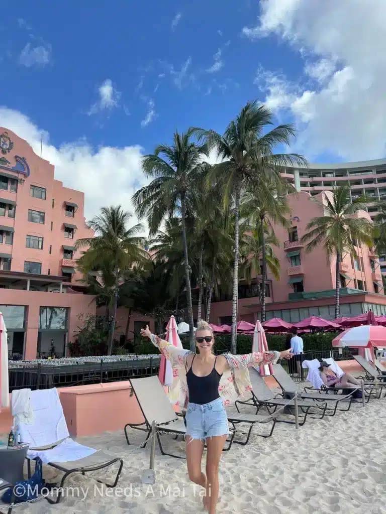 A woman smiling and standing with her arms outstretched on the beach and in front of the Royal Hawaiian Hotel in Waikiki, Oahu, Hawaii. 