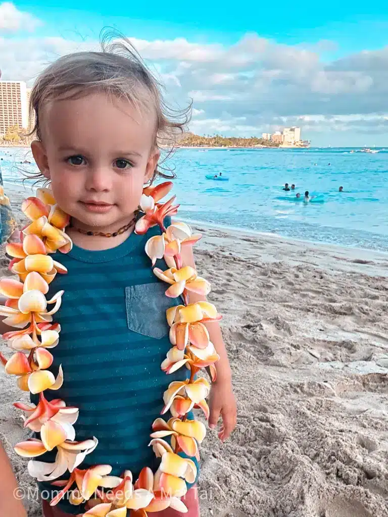 A toddler standing on the beach in Waikiki with a plumeria flower lei around his neck. 