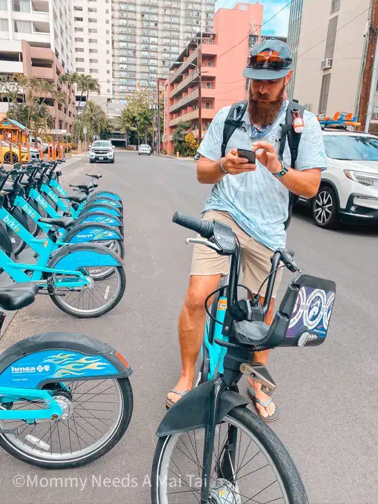 A man looking at directions on his phone while he is stopped and standing while on a Biki Bicycle Rental in Waikiki. 