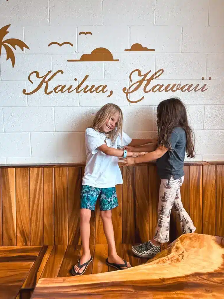 Two kids tickling each other and laughing in front of a wall that reads "Kailua, Hawaii."