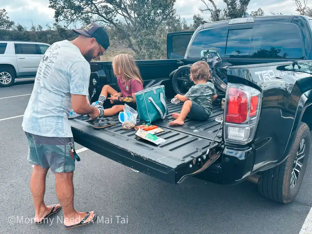 A dad putting shoes on a kid's foot as the young boy has a snack in the back of truck bed in Oahu, Hawaii. 