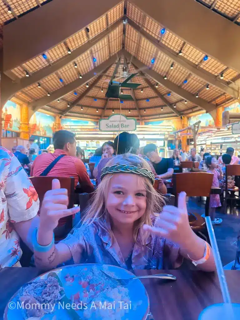 A boy giving two shakas at a luau with a headpiece on and a plate full of food.