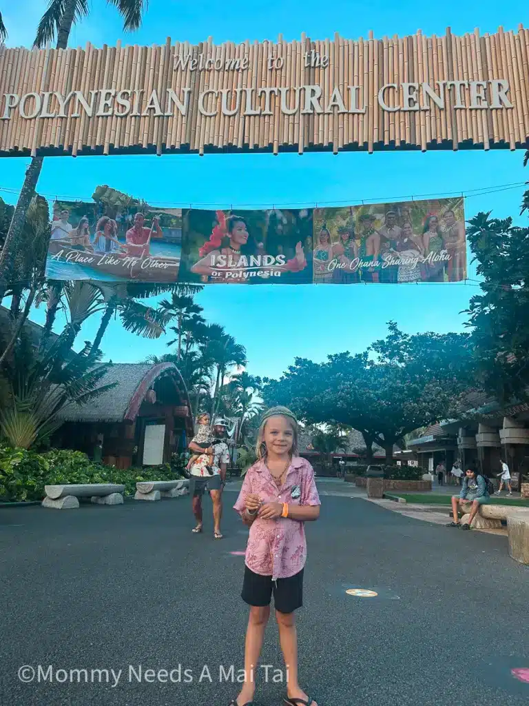 A young boy with long hair in an aloha shirt stands under a sign that reads "Polynesian Cultural Center."