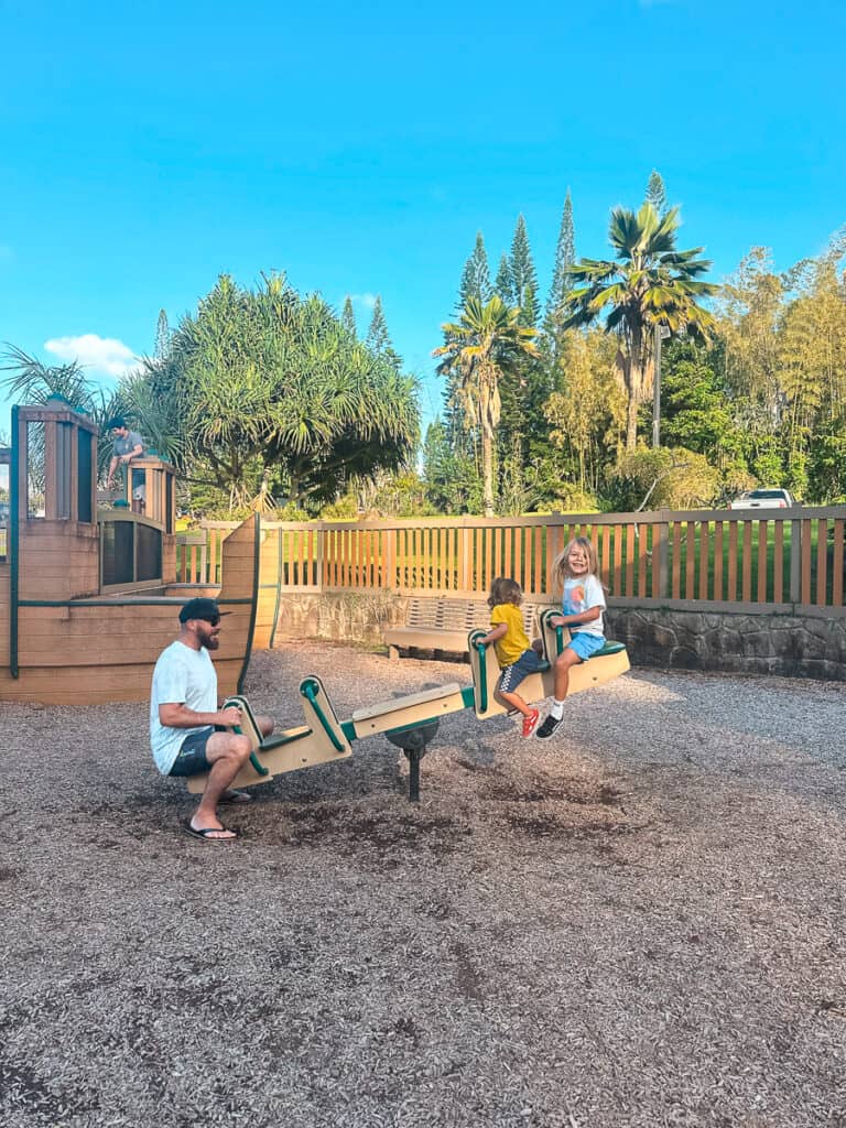 A man sits on a teeter-totter while two young kids are lifted into the air at a playground on Kauai. 