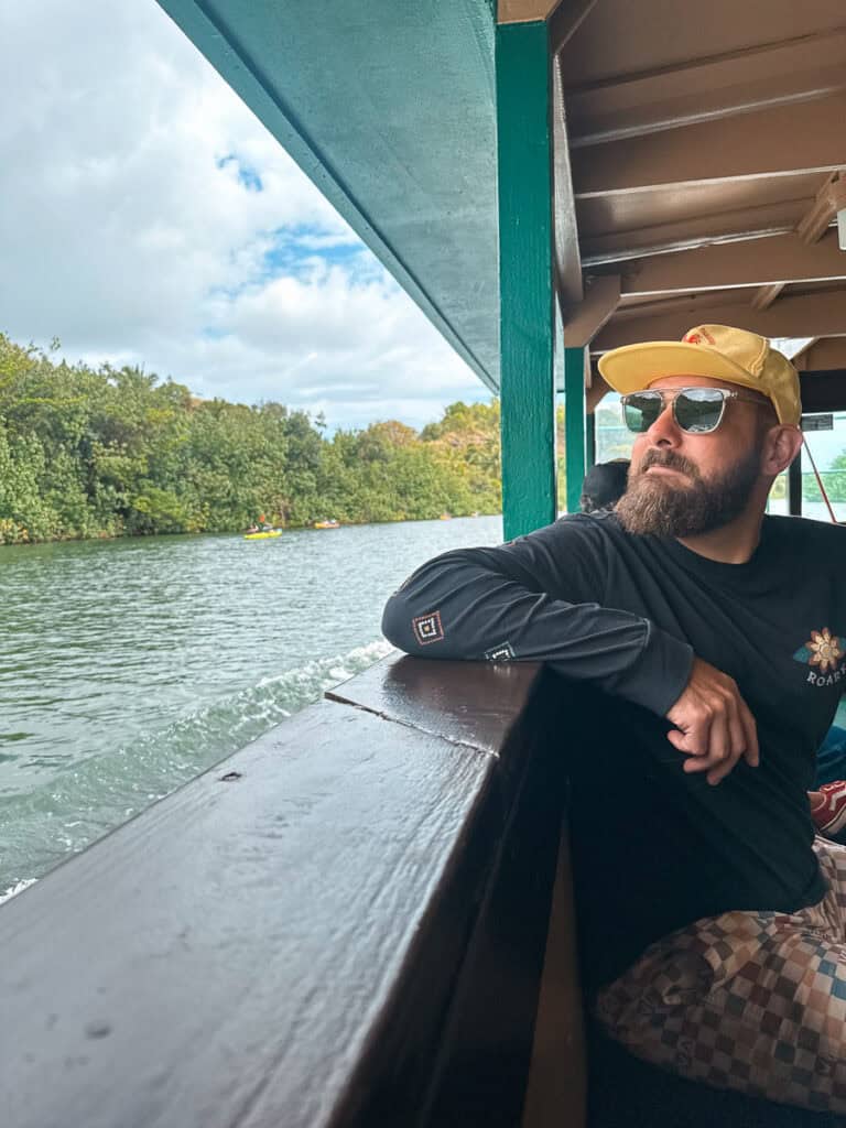 A man wearing a hat and sunglasses looks out at the Wailua River with green trees in the background. 