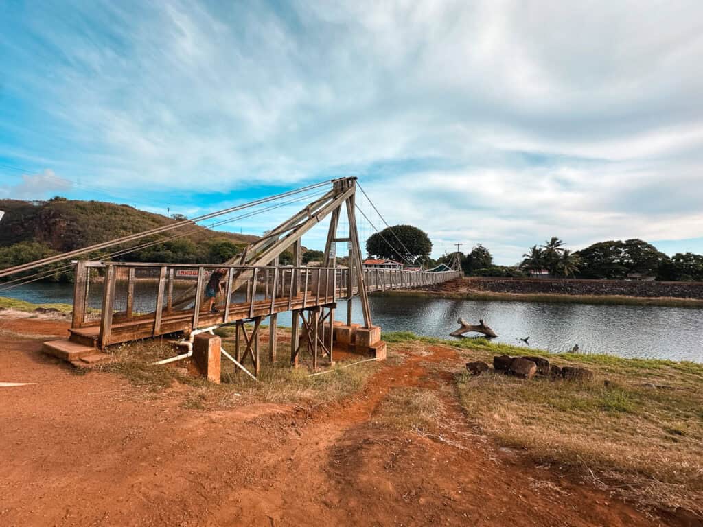 A wide view of the Hanapepe Swinging Bridge with water, trees, and cloud-laced blue skies. 