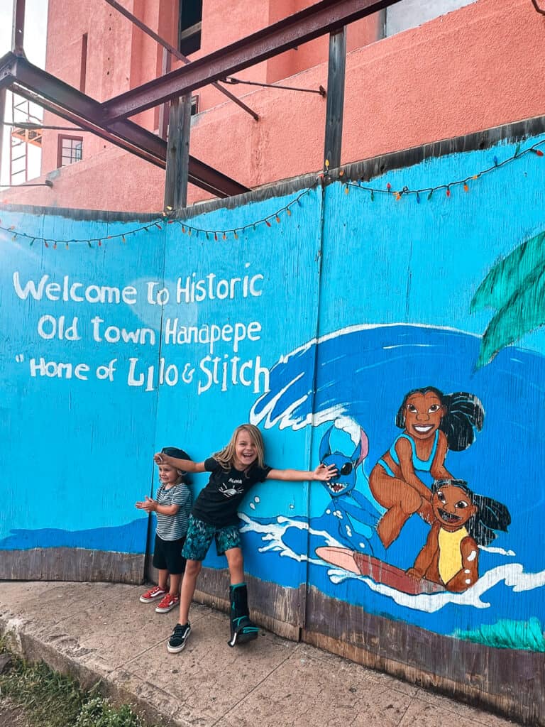 Two kids standing in front of mural that reads "Welcome to historic old town Hanapepe 'Home to Lilo & Stich'" on Kauai. 