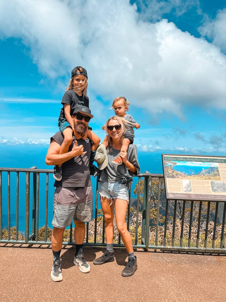 Parents exploring fun things to do on Kauai with kids, while standing and smiling with kids on their shoulders at Kalalau Valley Lookout at Koke'e State Park on Kauai.