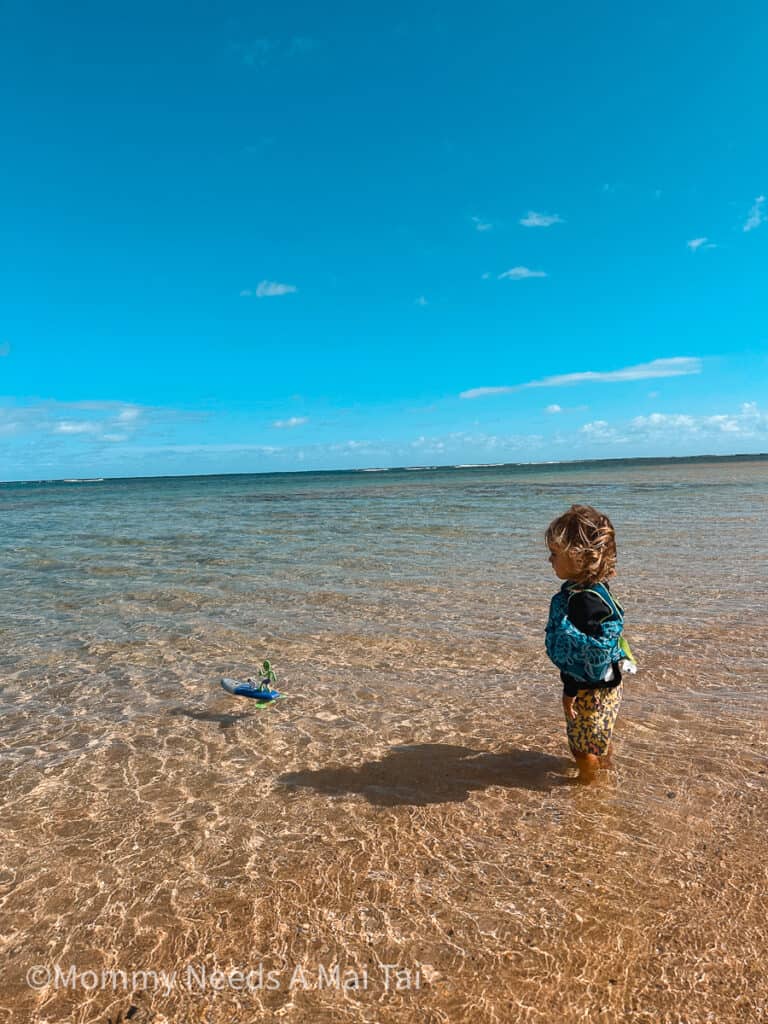 A young toddler wearing water wings looks out at the ocean while playing with a surf board toy.