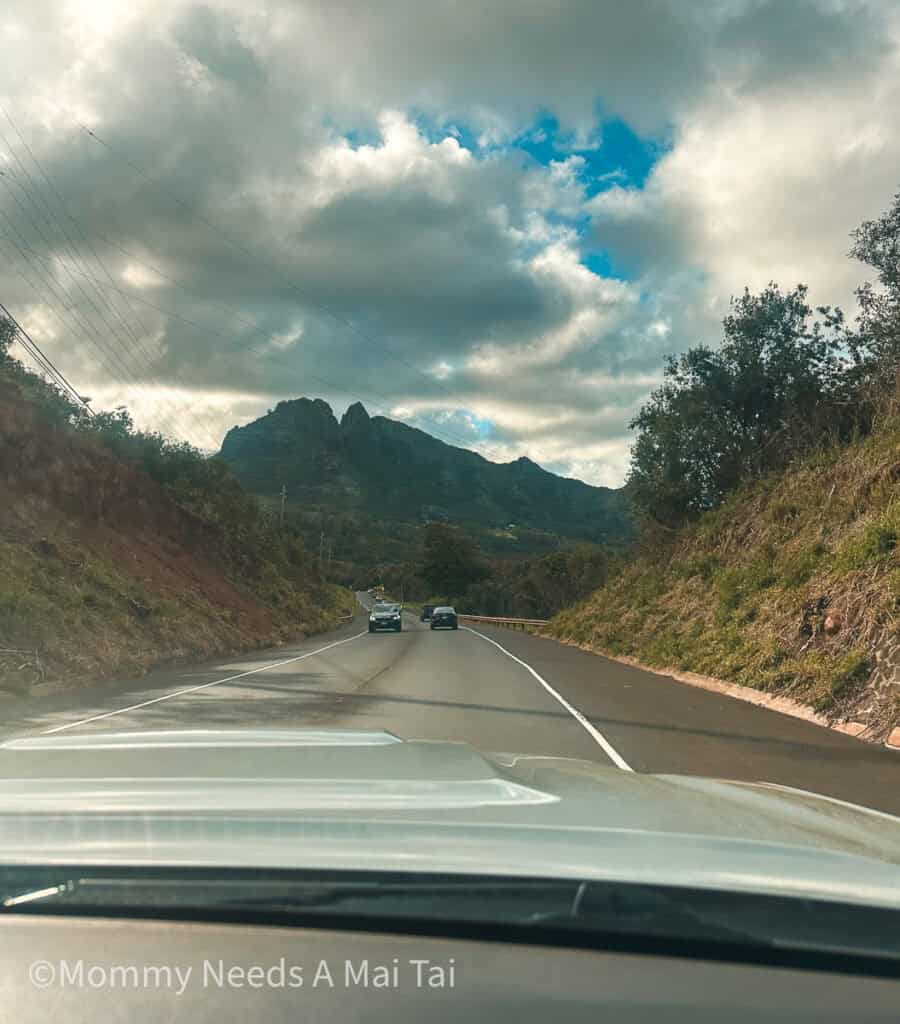 A long road in Kauai with lush mountains on each side of the car and jagged mountains up ahead. 