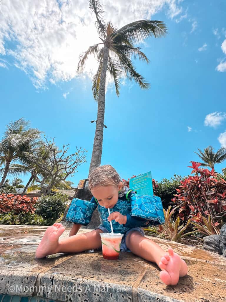 A young toddler wearing water wings drinks from a frozen drink by a resort pool on Kauai, with a palm tree behind him. 