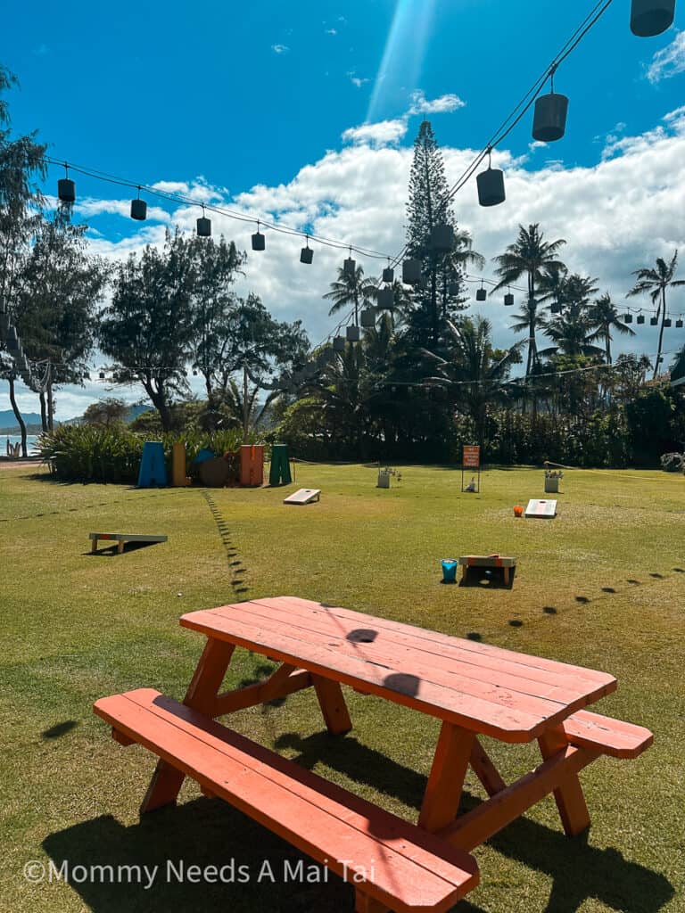 A view of Lava Lava Beach Club's outdoor play space, with picnic tables, bags game, and and aloha sign with the ocean in the background. 