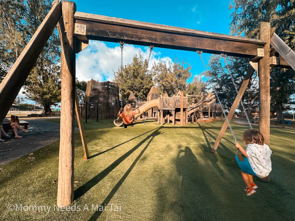 Two kids swinging on a wooden playground at Lydgate Beach Park in Kauai, Hawaii. 