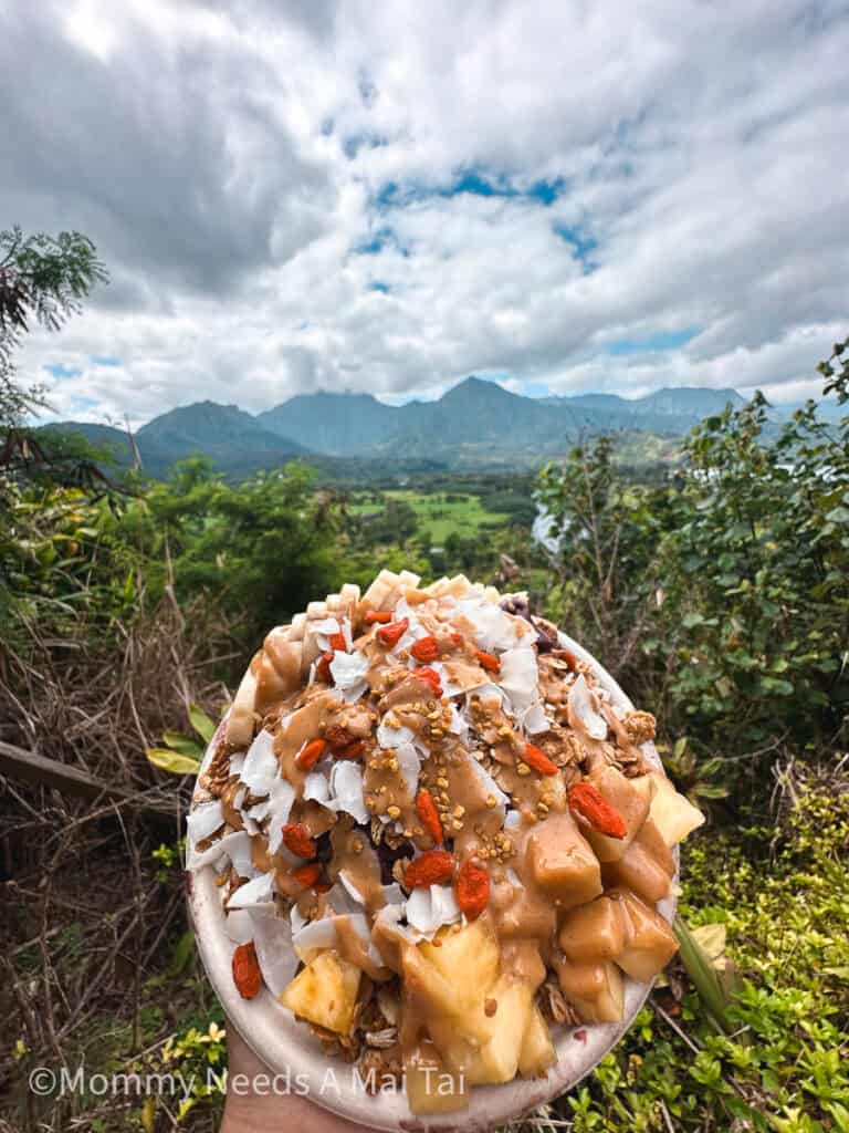 An Acai bowl topped with fresh pineapple and coconut being held up in front a view of the mountains at Nourish Hanalei on Kauai. 