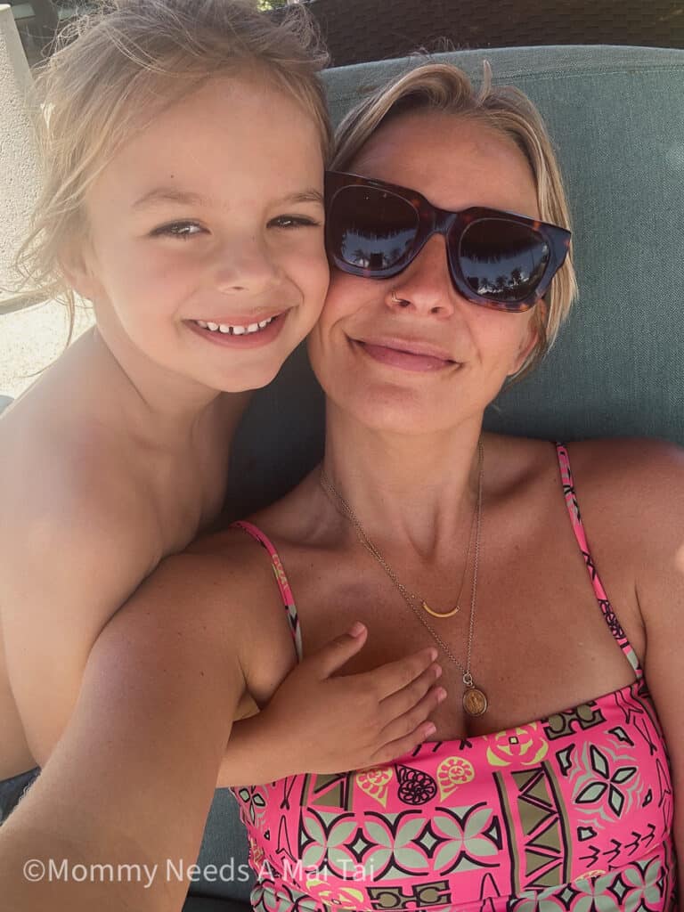 A young boy with long hair smiling and putting his hand on his mom, who is wearing sunglasses and lounging on chair at a resort pool.