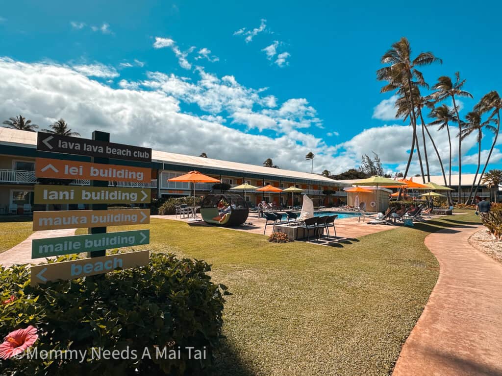 A wide view of Kauai Shores Hotel on Kauai, with signs telling you where to go, hotel pool with umbrellas, and palm trees blowing in the distance. 