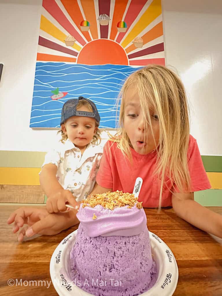 Two kids making excited faces as they look down at a purple shave ice with coconut toppings from Wailua Shave Ice on Kauai. 