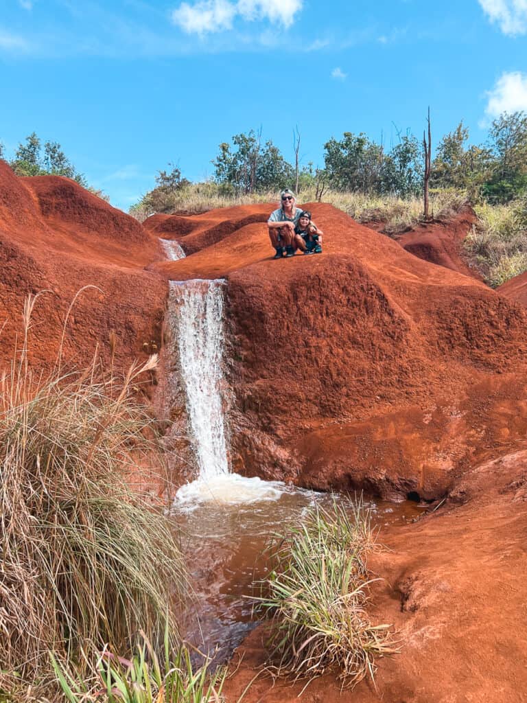 A mom and young boy sitting on top of a Red Dirt Falls mount next to a waterfall in Waimea Canyon, Kauai. 