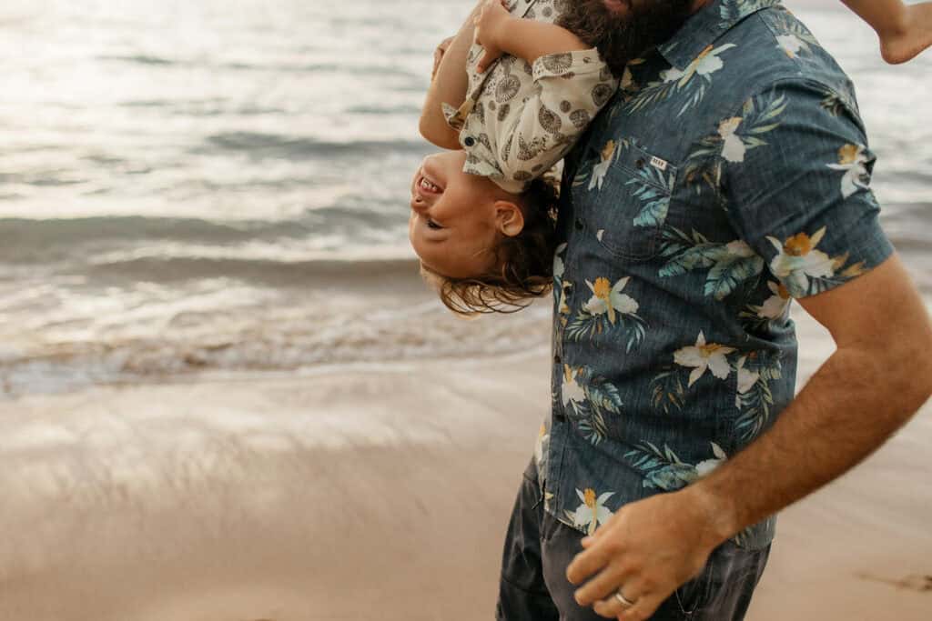 A toddler handing upside down from his dads arm on a beach in Hawaii.