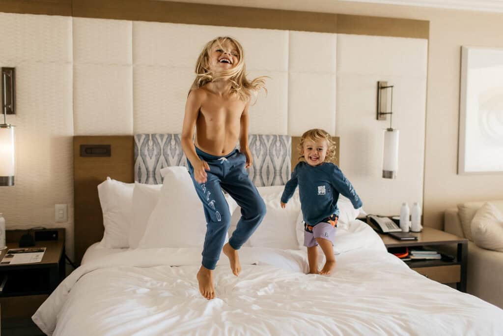 Two young children playing and jumping on a hotel bed.