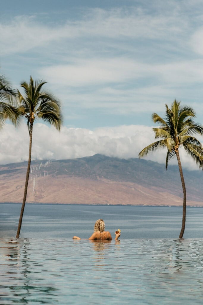 A woman looking out at the ocean in the infinity pool at Four Seasons Maui, with palm trees on each side.