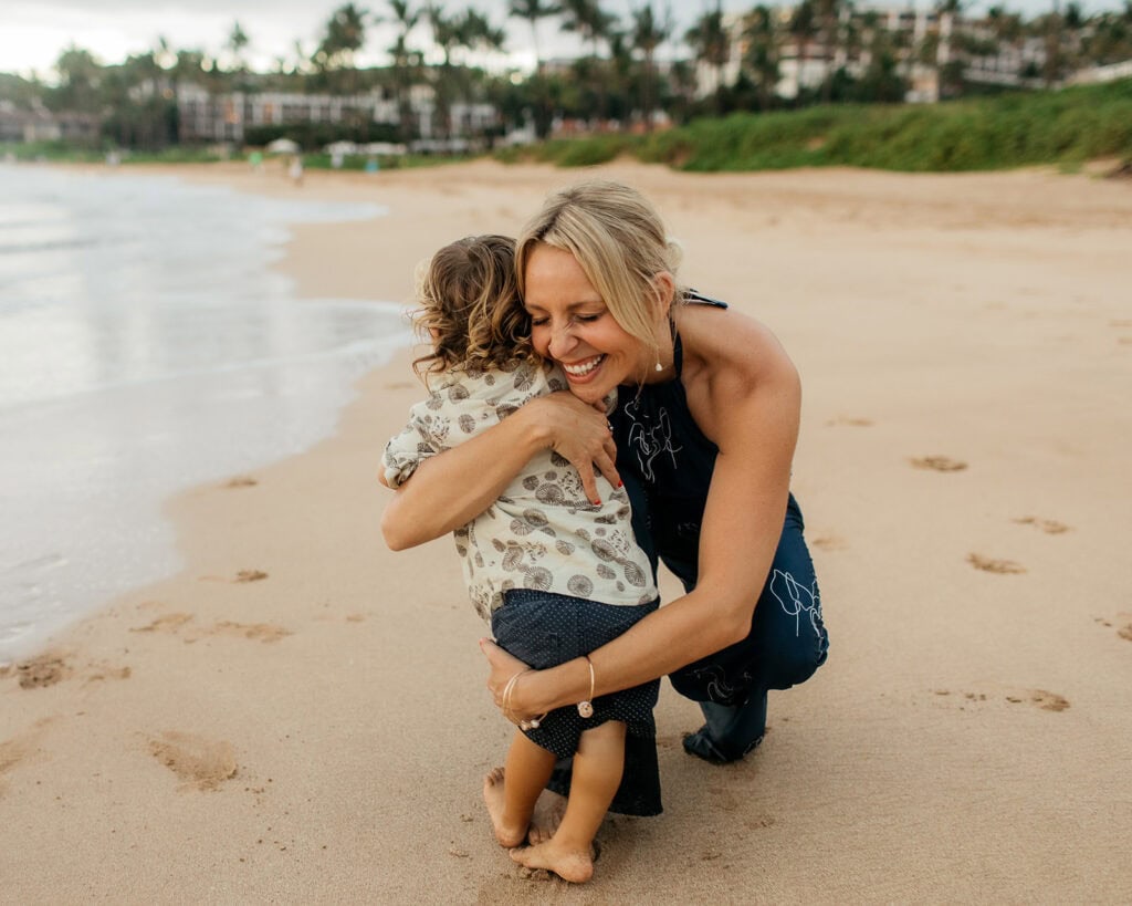 A mom hugging a toddler, barefoot on the beach in Hawaii.