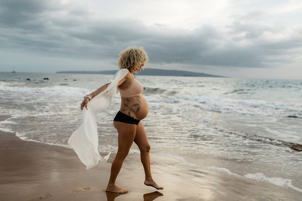 A pregnant woman in a swim suit with a cover up blowing in the wind on a beach in Hawaii.