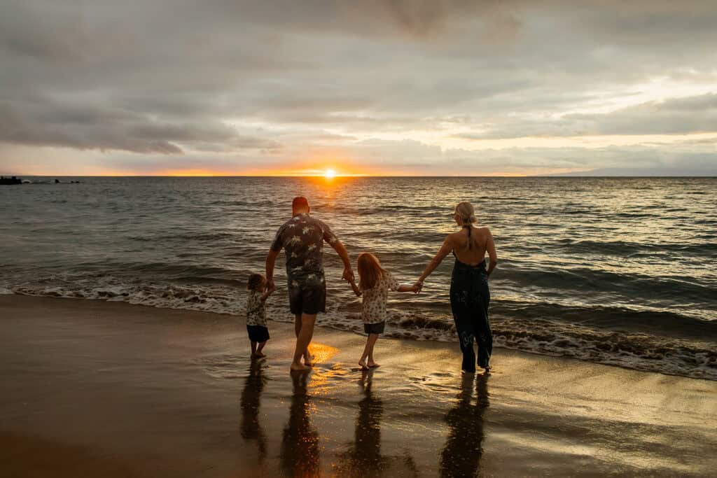 A family holding hands and looking out at the ocean while the sun sets on a beach in Hawaii.